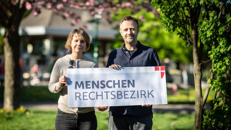 Bezirksvorsteherin Stefanie Lamp und Bezirksrad Stefan Jagsch halten gemeinsam ein Plakat in den Händen mit der Aufschrift "Menschenrechtsbezirk".
