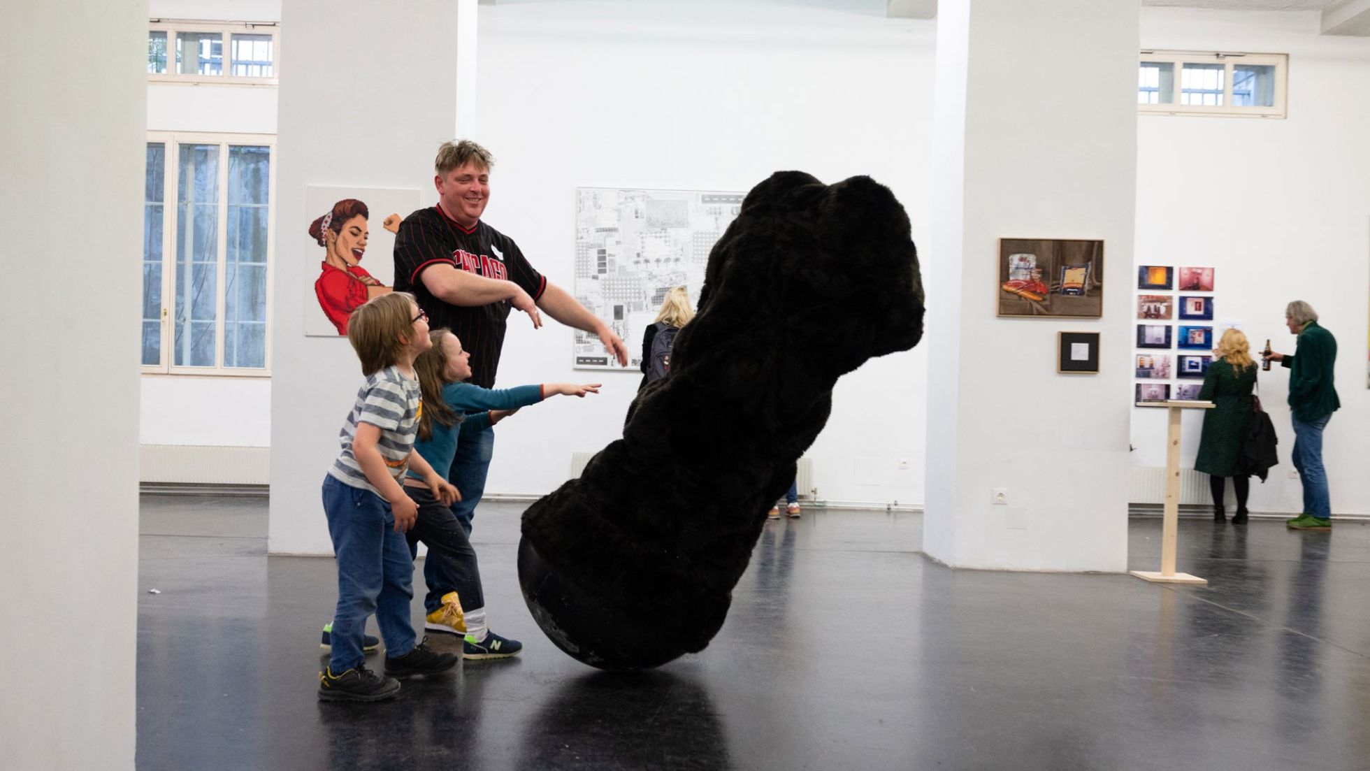 En Mann und 2 Kinder bewegen eine große, schwarze Skulptur, die mitten in einem Ausstellungsraum steht