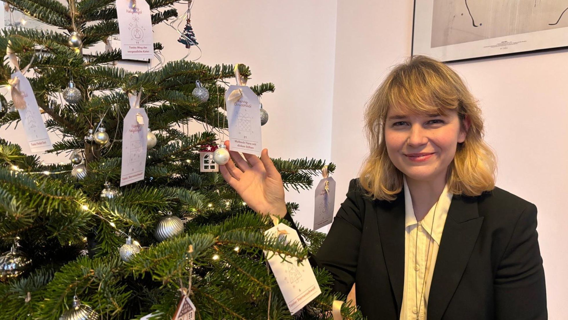 Eine Frau steht neben einem Christbaum, auf dem neben Kugeln auch verschiedene Wunschzettel hängen.