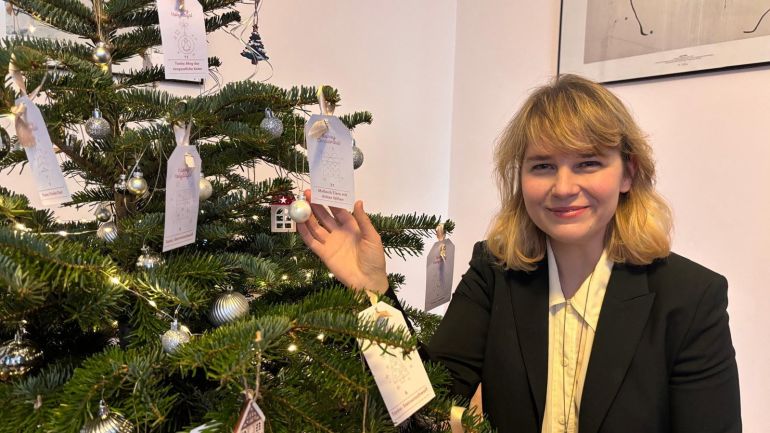 Eine Frau steht neben einem Christbaum, auf dem neben Kugeln auch verschiedene Wunschzettel hängen.