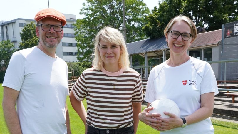 1 Mann und 2 Frauen stehen fröhlich auf einem grünen Sportrasen.