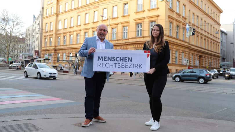 2 Personen halten ein Schild mit "Menschenrechtsbezirk" an einer Kreuzung vor einem historischen Gebäude