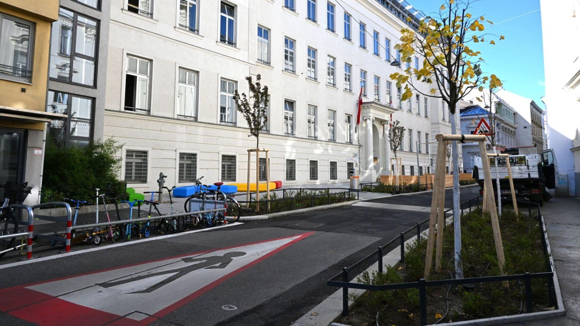 Neuer Schulvorplatz mit bunten Sitzmöbeln, Beeten und Bäumen, davor eine Straßenmarkierung für "Achtung Kinder queren".