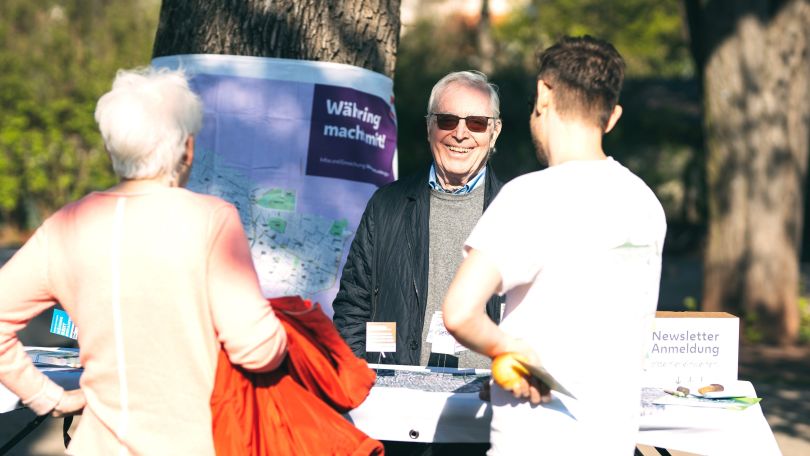 copyright: epilogy.photography/Tim Dornaus 3 Personen sind fröhlich in einem Gespräch an einen Infotisch im Freien.