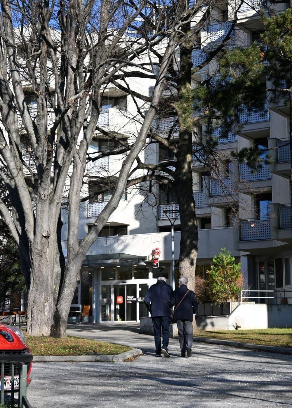 2 Senioren auf einem Weg vor einem Wohngebäude.
