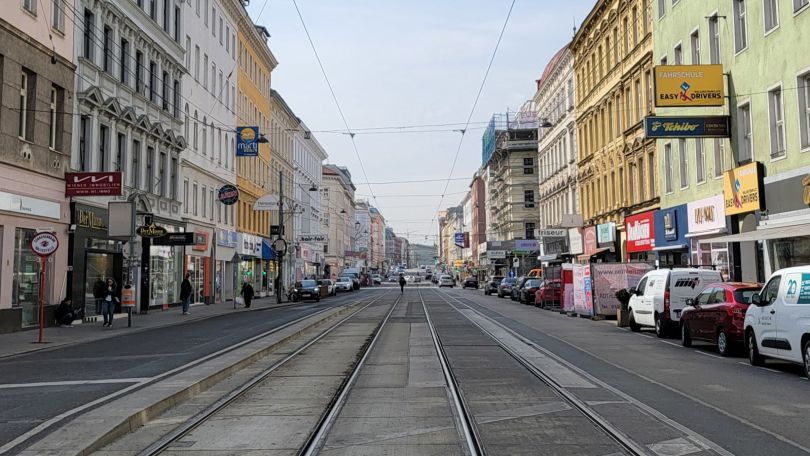 Blick in eine Straße mit Fahrbahn, Schienen, an der Seite parkende Autos und Hausfassaden