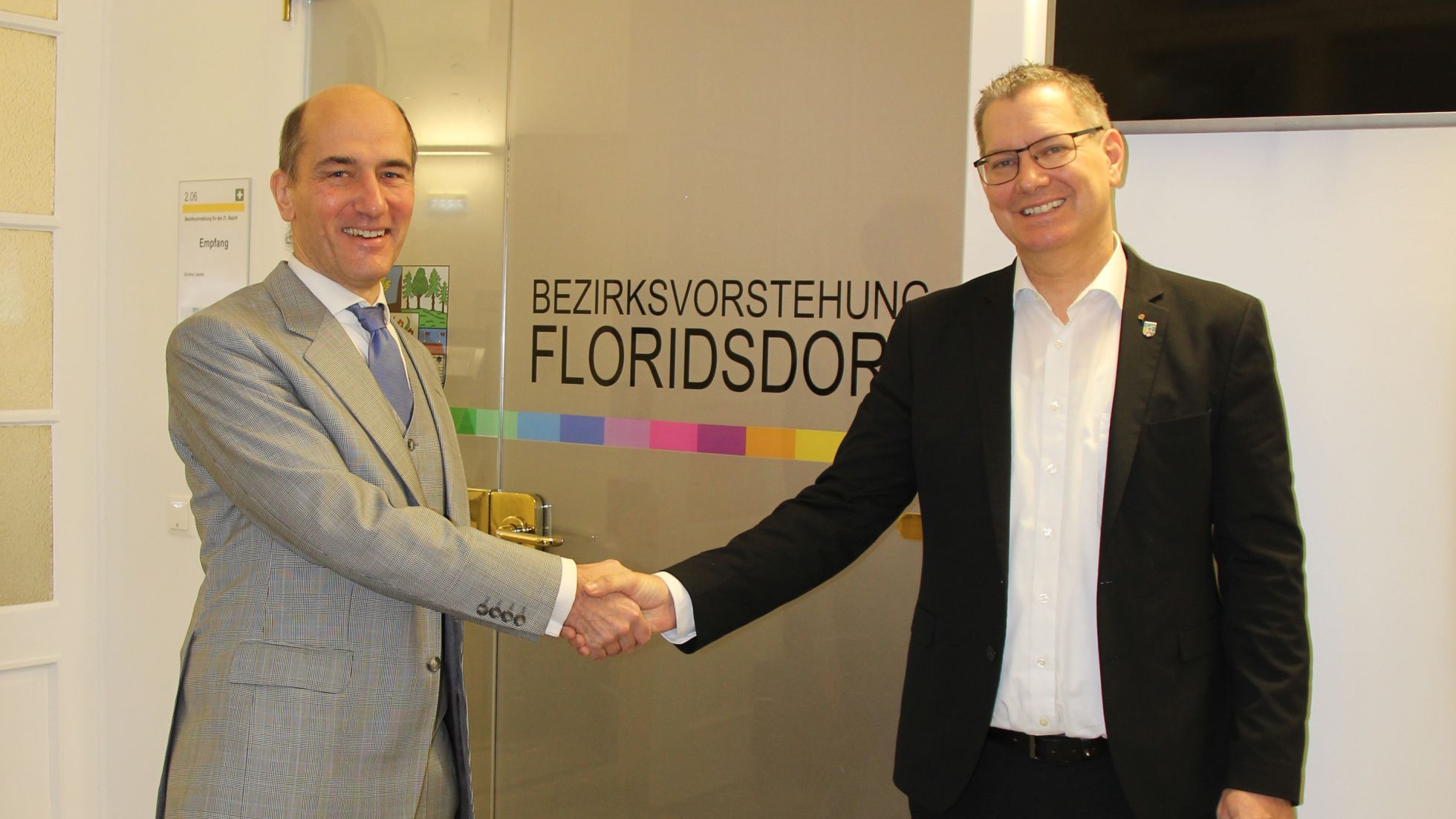 2 Herren in Anzug schütteln Hände vor Tür mit Aufschrift "Bezirksvorstehung"