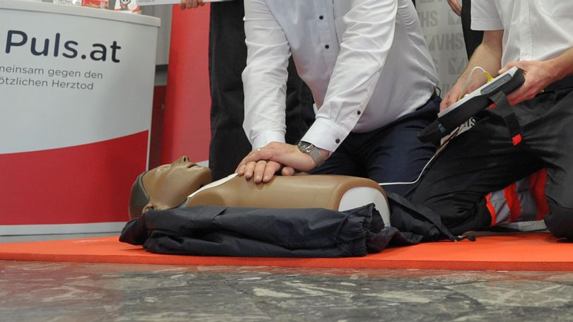 Reanimationspuppe liegt am Boden und 2 Menschen üben damit die Herzdruckmassage und das Anhängen eines Defibrillators