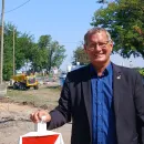 Bezirksvorsteher Georg Papai stützt sich auf ein Baustellenzeichen, dahinter sind ein Bus, zwei Bagger und ein Kipplaster neben einer Straße und Bäumen