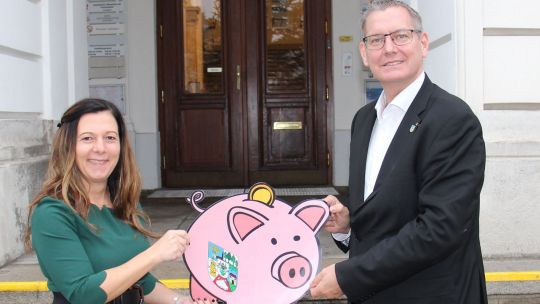 Eine Frau und ein Mann stehen vor einem Amtsgebäude und halten ein Sparschwein aus Karton zwischen sich