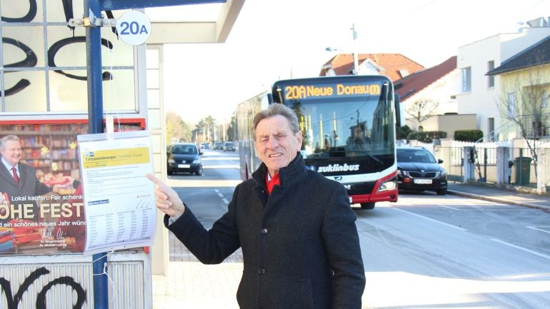 Mann in schwarzem Mantel zeigt auf den Fahrplanaushang bei der Bushaltestelle, im Hintergrund ein Linienbus.