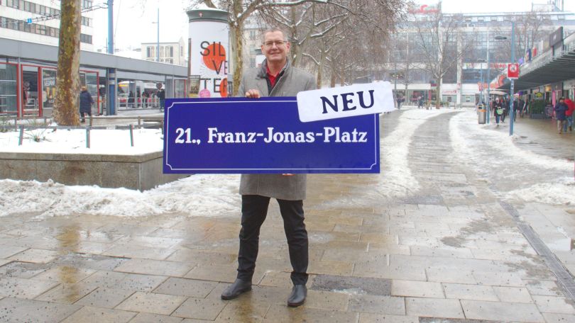 Ein Mann hält ein vergrößertes Straßenschild "21., Franz-Jonas-Platz" hoch.