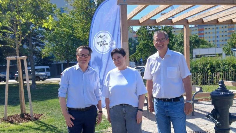 2 Politiker und eine Politikerin im neuen Park neben Trinkbrunnen und Holzpergola.