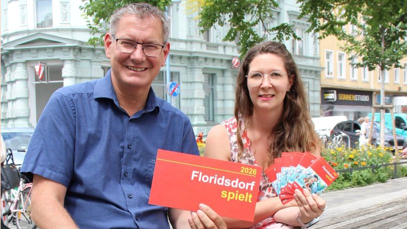 copyright: BV 21 Bezirksvorsteher Georg Papai und Bezirksrätin Katharina Allerbauer sitzen nebeneinander auf einer Parkbank und halten das Programm zum Ferienspiel in den Händen
