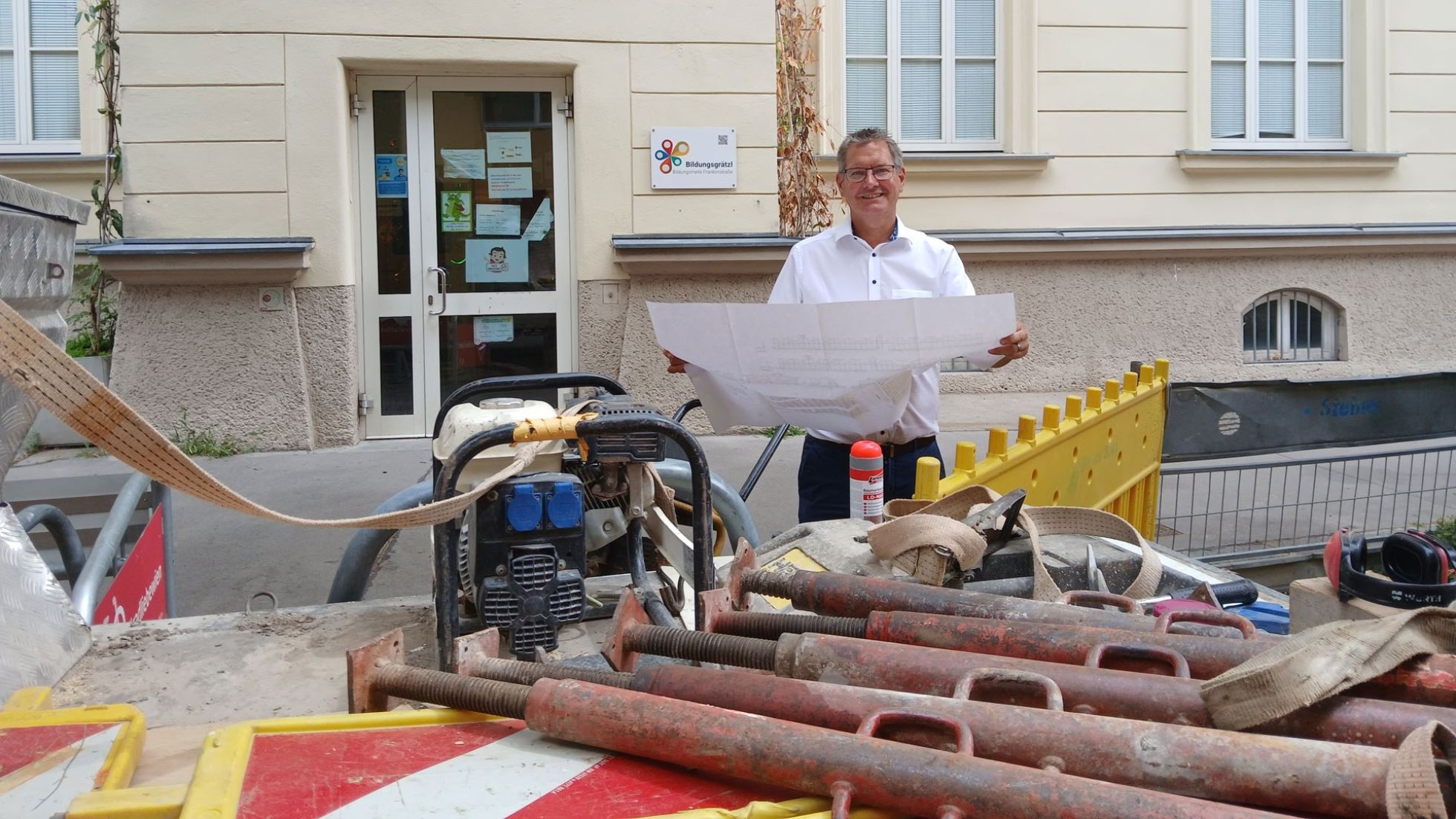 Bezirksvorsteher Papai steht bei der Baustelle Schulvorplatz Mengergasse und hält einen Plan in den Händen