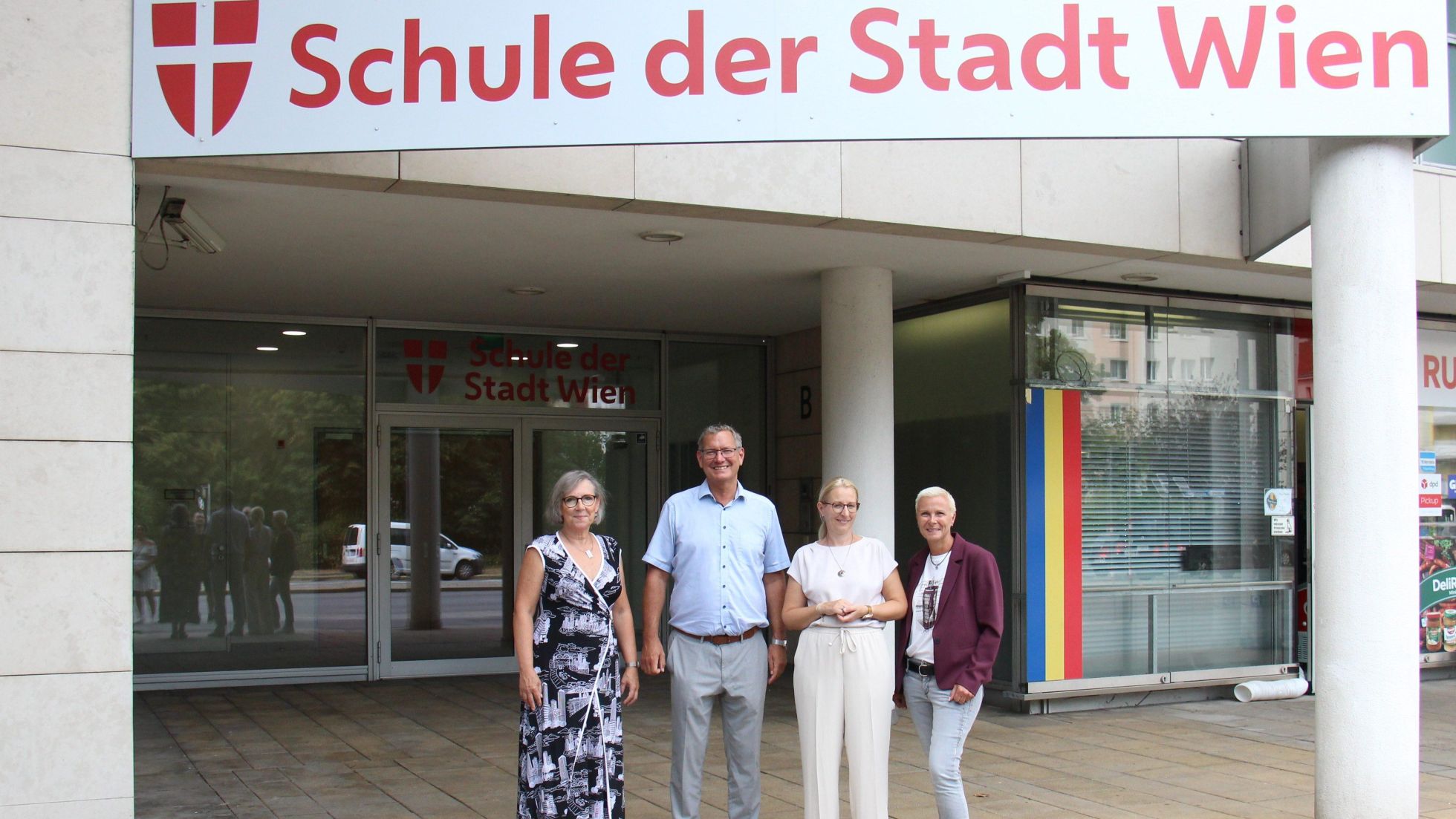 copyright: BV 21 Georg Papai, Andrea Trattnig, die Schuldirektorin und die Schulqualitätsmanagerin stehen vor dem Eingang. Darüber prangt ein großes Schild "Schule der Stadt Wien".