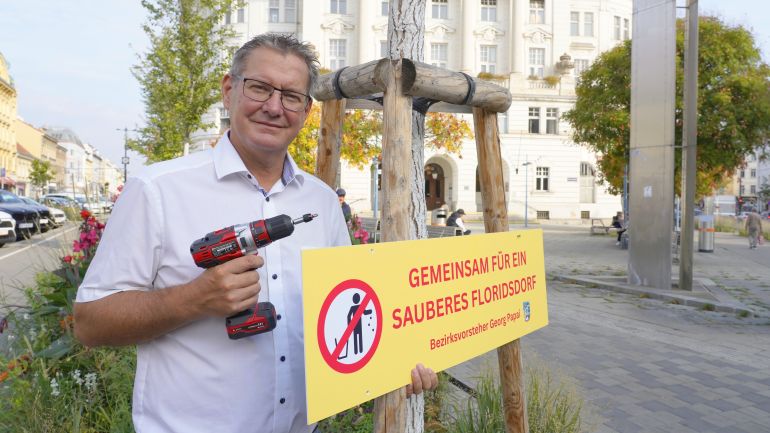 copyright: BV 21 Ein Mann mit einem Bohrer in der rechten Hand und einem Schild mit der Aufschrift "Gemeinsam für ein sauberes Floridsdorf" in der linken Hand.