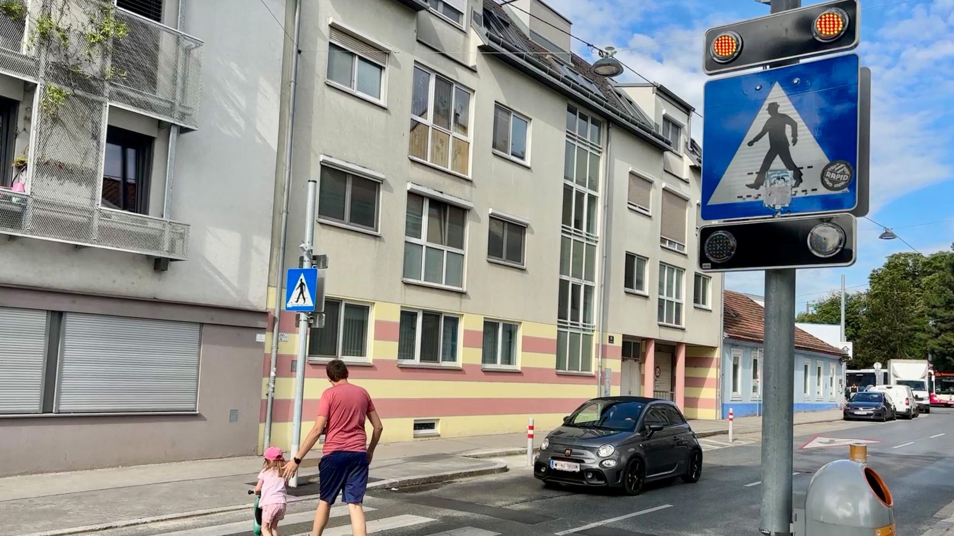 Ein Mann geht mit einem kleinen Kind am Roller über einen Zebrastreifen. Ein Auto steht davor. Das Verkehrsschild "Fußgängerübergang" ist mit orangen Blinklichtern ausgestattet.