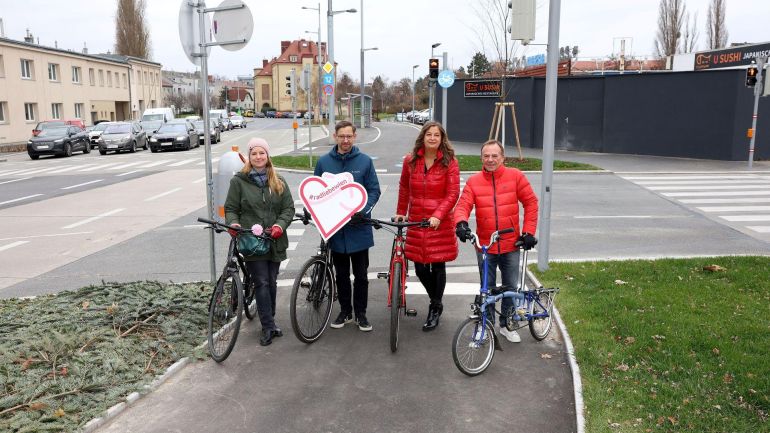 4 Menschen stehen mit Fahrrädern auf einem breiten Radweg vor einer Kreuzung.