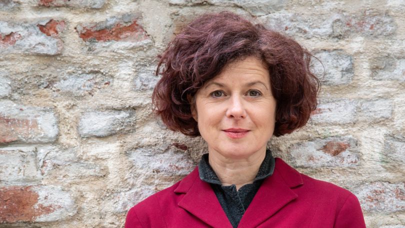 Frau mit lockigen braunen Haaren und rotem Blazer steht vor einer Backsteinmauer mit verschränkten Händen.