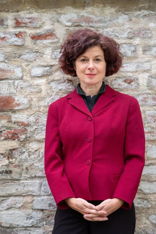 Frau mit kurzen braunen Locken und rotem Blazer verschränkt die Hände vor der Hüfte