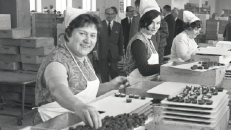 Schwarz-weiß-Aufnahme von Frauen, die Konfitüre verpacken. Im Hintergrund Stapel mit fertigen Packungen sowie eine Gruppe von Männern in Anzügen