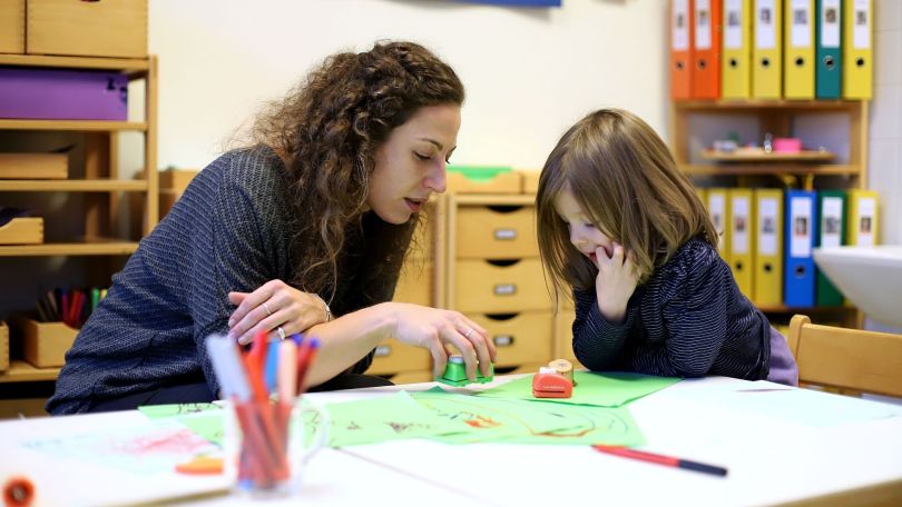copyright: MA 10 - Wiener Kindergärten/VOTAVA Frau mit langen braunen Locken sitzt bei einem kleinen Mädchen an einem Tisch, darauf bunte Zettel und Buntstifte.
