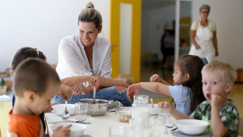 copyright: MA 10 - Wiener Kindergärten/VOTAVA Kinder sitzen bei Tisch und essen aus Schüsseln, daneben eine junge, lächelnde Frau mit blonden, zusammengebundenen Haaren.