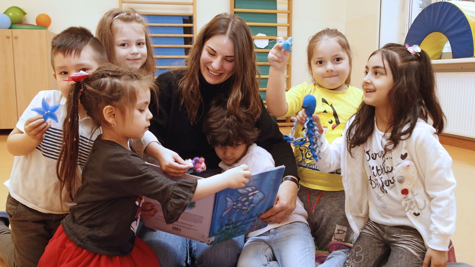 copyright: MA 10 - Wiener Kindergärten Kindergartenkinder sitzen rund um eine junge lächelnde Frau und schauen ein Buch an.