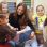 Kindergartenkinder sitzen rund um eine junge lächelnde Frau und schauen ein Buch an.