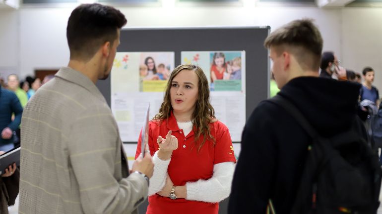 Frau bei Infostand erklärt 2 Interessenten etwas