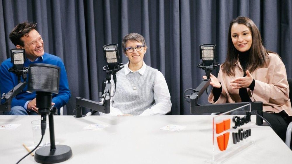 Ein Mann und zwei Frauen sitzen vor Podcast-Mikrofonen an einem Tisch.