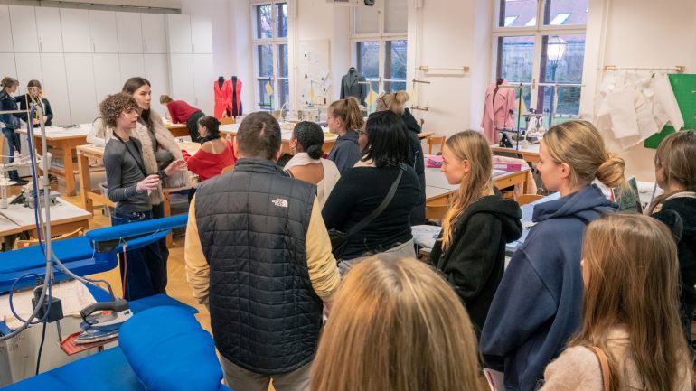 copyright: Modeschule Hetzendorf – Julian Vilim Gruppe von Schülern, die in einer Klasse mit Tischen und Nähmaschinen etwas gezeigt bekommen