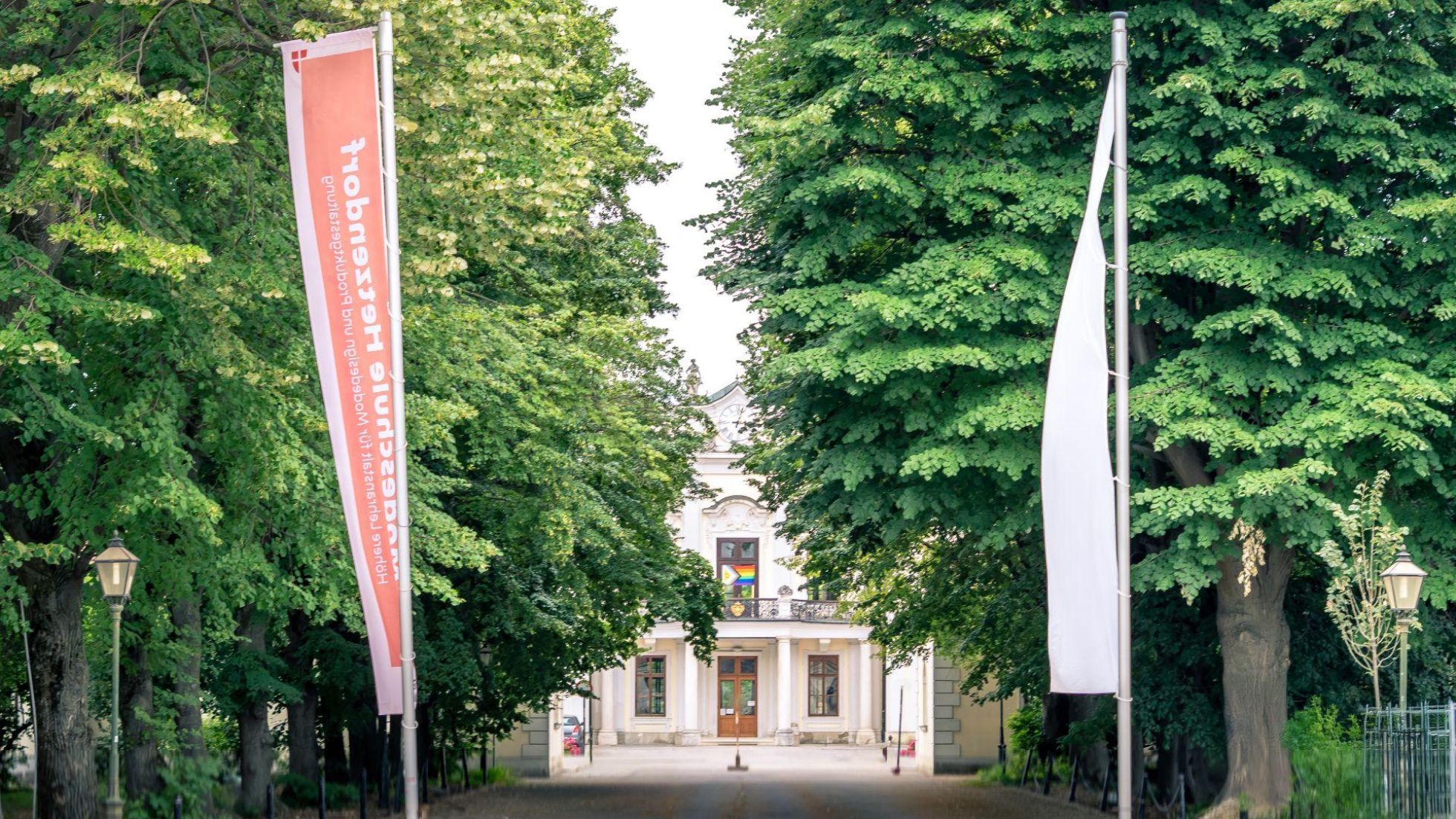 copyright: Modeschule Hetzendorf Eine rote und eine weiße Fahne vor einer Baumallee, dahinter das Schloss Hetzendorf.