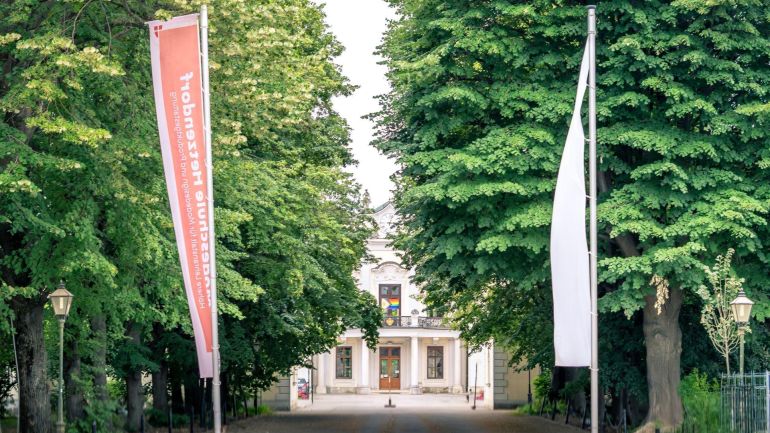 copyright: Modeschule Hetzendorf Eine rote und eine weiße Fahne vor einer Baumallee, dahinter das Schloss Hetzendorf.