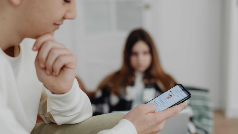 Junge schaut aufs Handy, im Hintergrund ein Mädchen