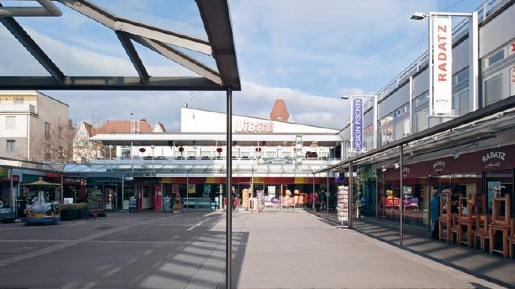 befestigter Freibereich, umgeben von Gebäuden mit Geschäften im Erdgeschoß und Glasvordächern