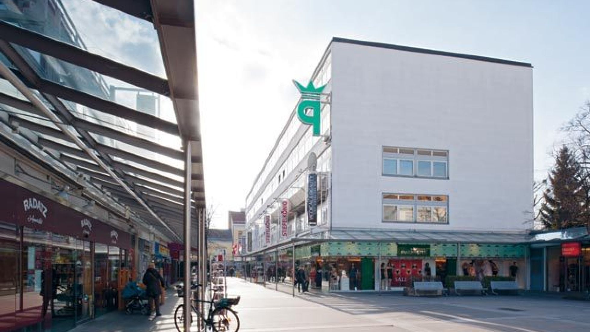 Blick unter einem Glasvordach entlang eines länglichen Gebäudes, rechts parallel dazu ein weiteres Gebäude