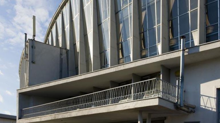 Fassade aus Glas zwischen Betonstützen, auf darunter querliegendem Balkon, außen angebrachte Rohre