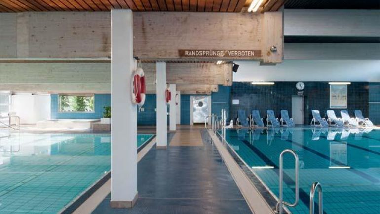 copyright: Fotografin: Pez Hejduk Innenbereich, links und rechts Teile von Schwimmbecken, im Hintergrund Liegestühle, oben massive Holzträger unter der Decke