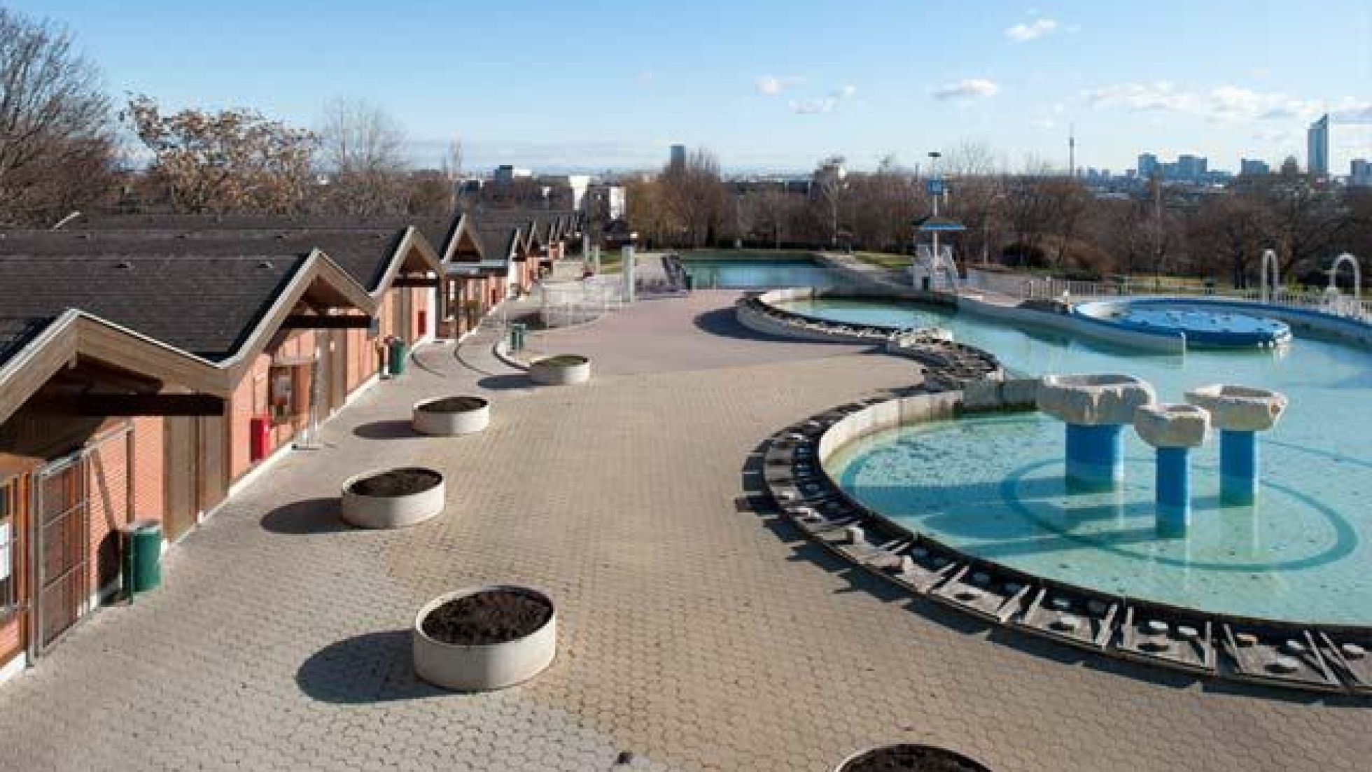 Außenbereich, gepflasterte Fläche, rechts ein Schwimmbecken in geschwungener Form, links Reihe von Holzhütten