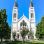 copyright: Stadt Wien / Christian Fürthner Kirche mit Zwillingstürmen, rund herum viele hohe Bäume und Wiese.