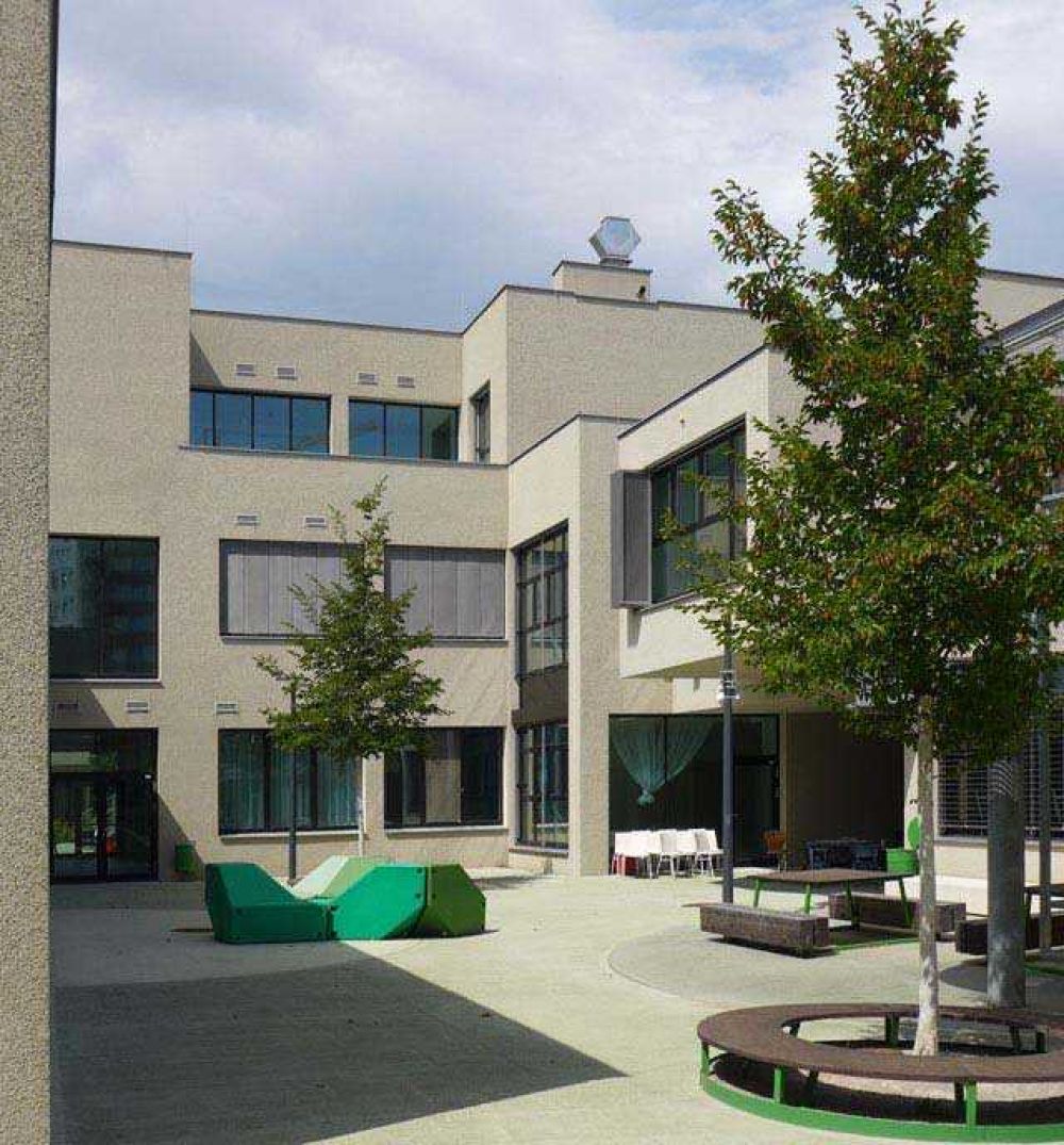 <p>Innenhof mit Sitzgelegenheiten</p> Innenhof mit grünen Kunststoffbänken, Holztischen und Stühlen. Den Verweilbereichen sind jeweils Bäume zur Verschattung zugeordnet.