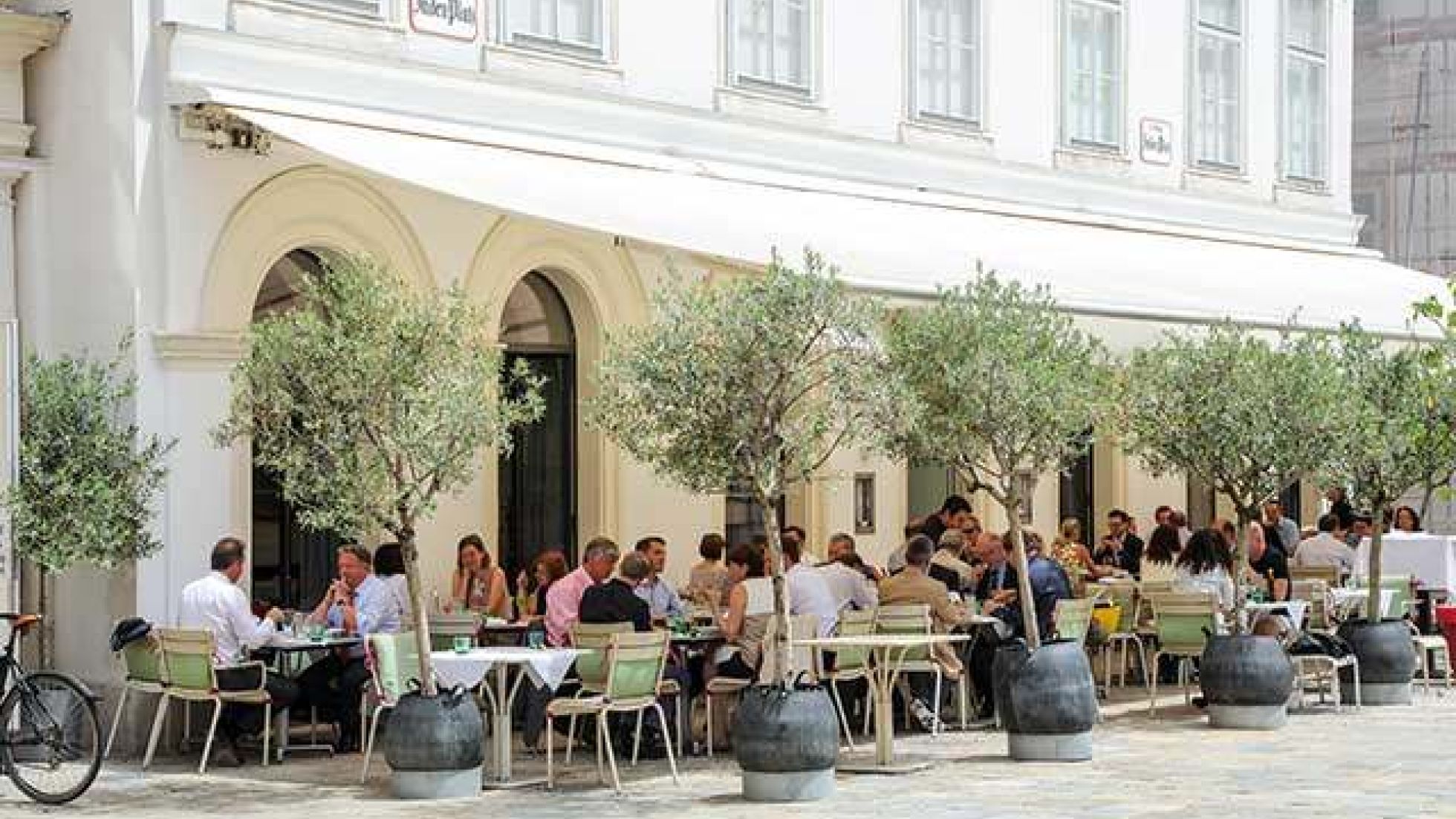 copyright: ©christian fürthner/MA19 Menschen sitzen an Tisch-Sessel-Kombinationen unter einer Markise, umgeben von Pflanztrögen mit Bäumen.