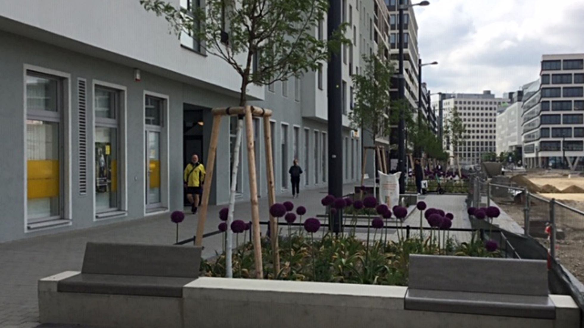 Wohngebäude, daneben Gehweg, Blumenbeet mit kleinen Bäumen und Bau-Kran