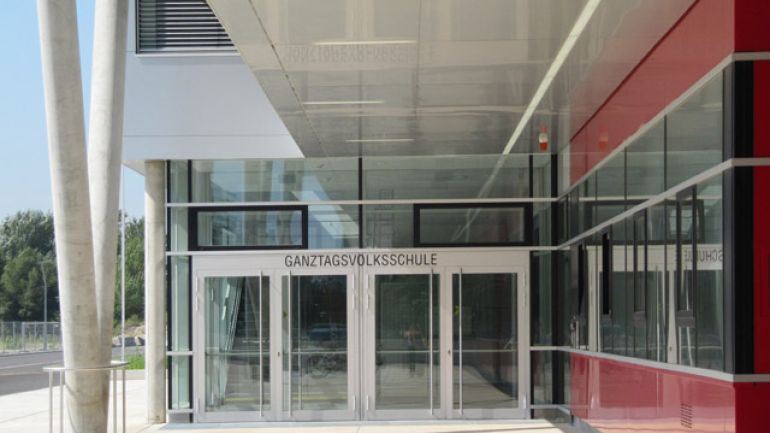 gedeckter Eingang der Ganztagsvolksschule, links vorgelagerte V-Stützenkonstruktion, rechts rote Fassade