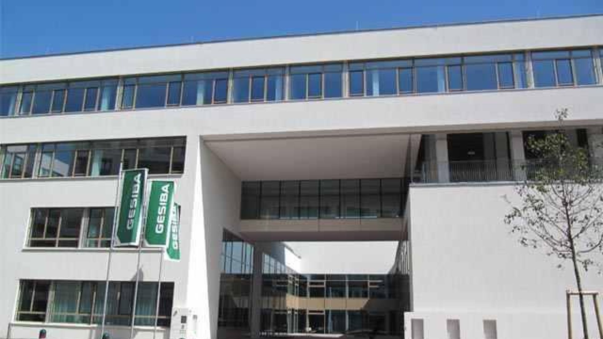 Frontalansicht des 4-geschoßigen, weißen Baukörpers mit Fensterbändern. Über einen 3-geschoßigen Ausschnitt sieht man in einen Innenhof.