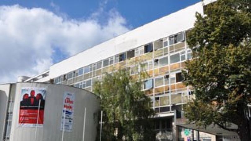 langgestrecktes Gebäude mit Fassade aus Fensterbändern, links im Vordergrund ein zylindrisches Gebäude