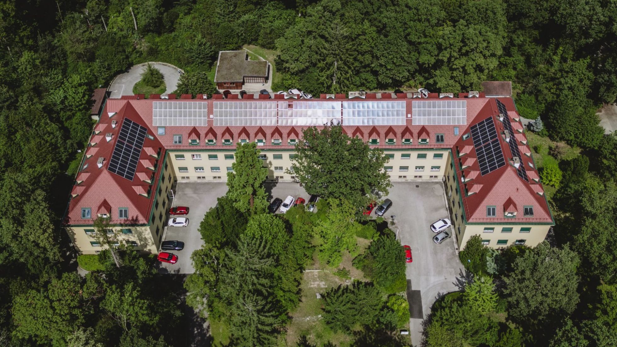Blick von oben auf ein Gebäude mit Parkplatz und Photovoltaikanlage auf dem Dach, umgeben von Wald.