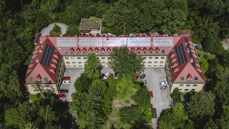 Blick von oben auf ein Gebäude mit Parkplatz und Photovoltaikanlage auf dem Dach, umgeben von Wald.